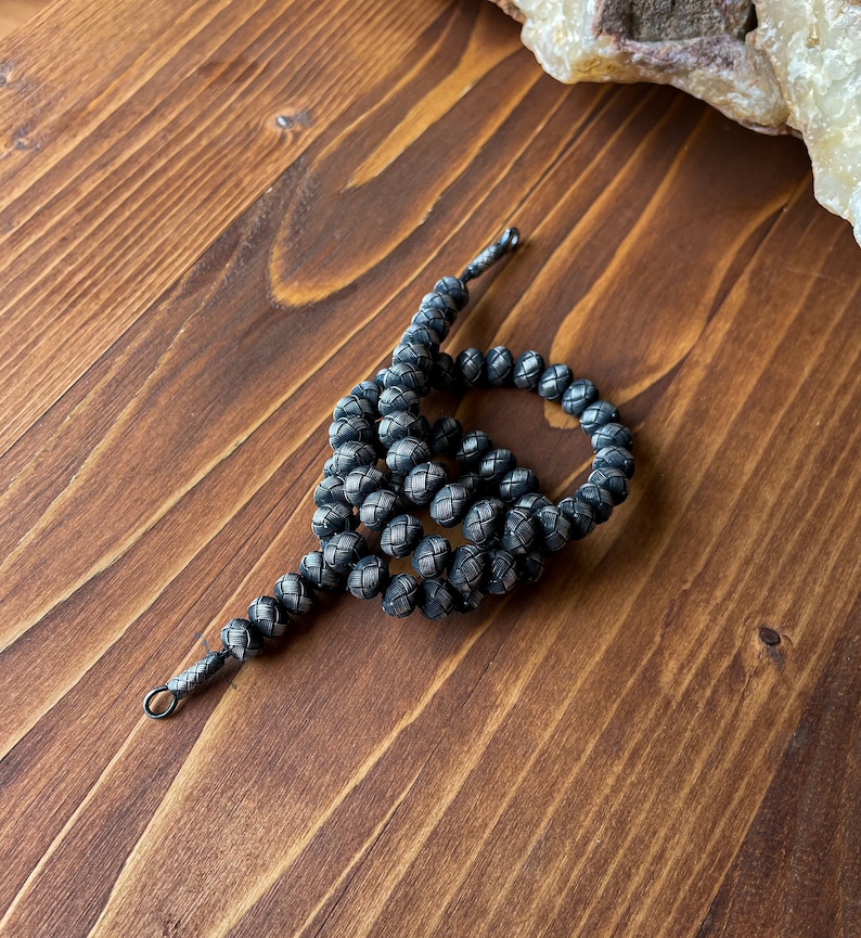 May include: A dark gray beaded necklace with a woven pattern. The beads are round and connected by a dark gray cord. The necklace is lying on a wooden surface.