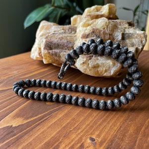 May include: A silver-toned beaded necklace with a woven pattern. The necklace is displayed on a wooden surface, with a light-colored, textured stone in the background. The beads are round and evenly spaced.