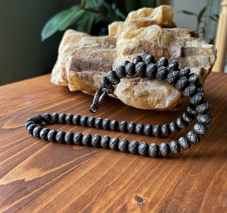 May include: A silver-toned beaded necklace with a woven pattern. The necklace is displayed on a wooden surface, with a light-colored, textured stone in the background. The beads are round and evenly spaced.