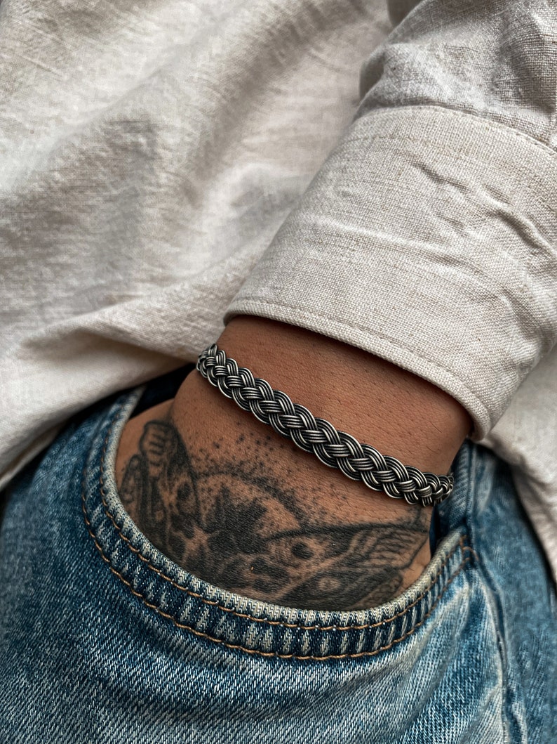 May include: A silver braided bracelet with a woven design. The bracelet is worn on a person's wrist.