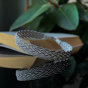 Op de afbeelding: Een zilveren gevlochten armband met een geweven ontwerp. De armband ligt op een lichtbruin oppervlak.