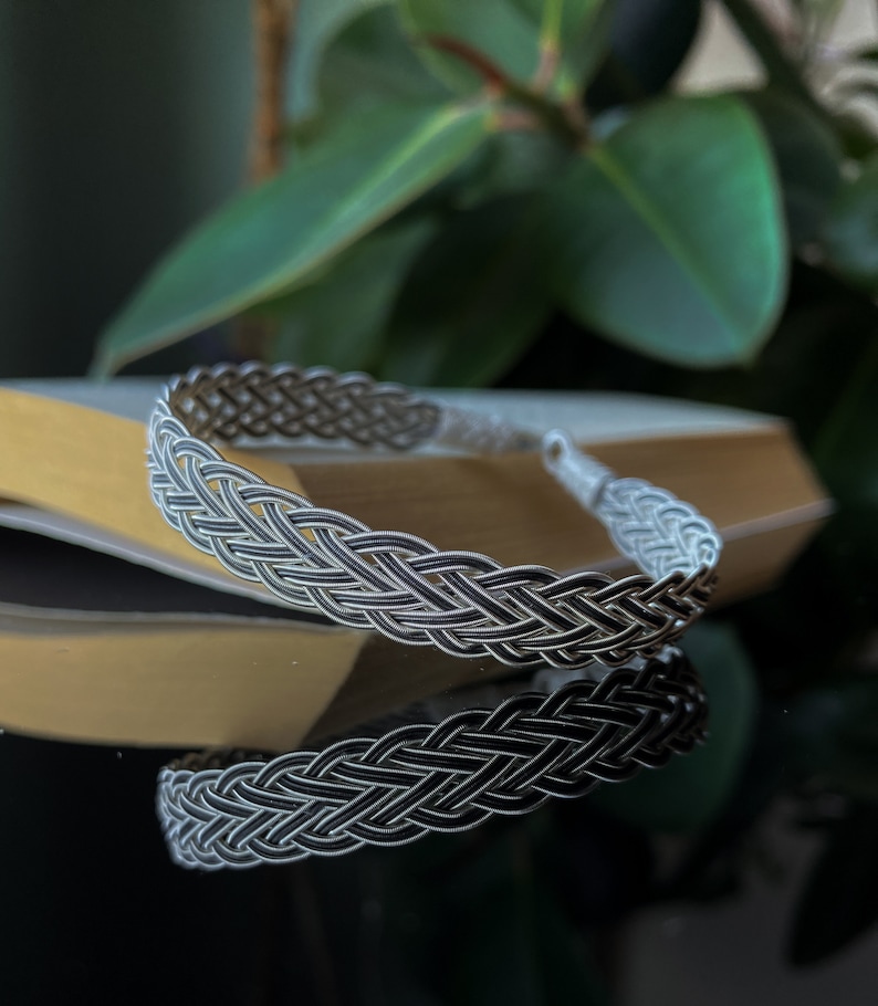 Op de afbeelding: Een zilveren gevlochten armband met een geweven ontwerp. De armband ligt op een lichtbruin oppervlak.