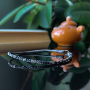 May include: A black braided chain necklace with a silver clasp. The necklace is lying on a reflective surface.
