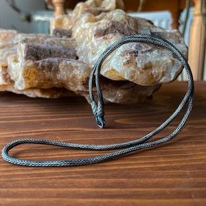 Pode incluir: Um colar de corrente de prata com um design tran&ccedil;ado e um fecho. O colar est&aacute; sobre uma superf&iacute;cie de madeira.