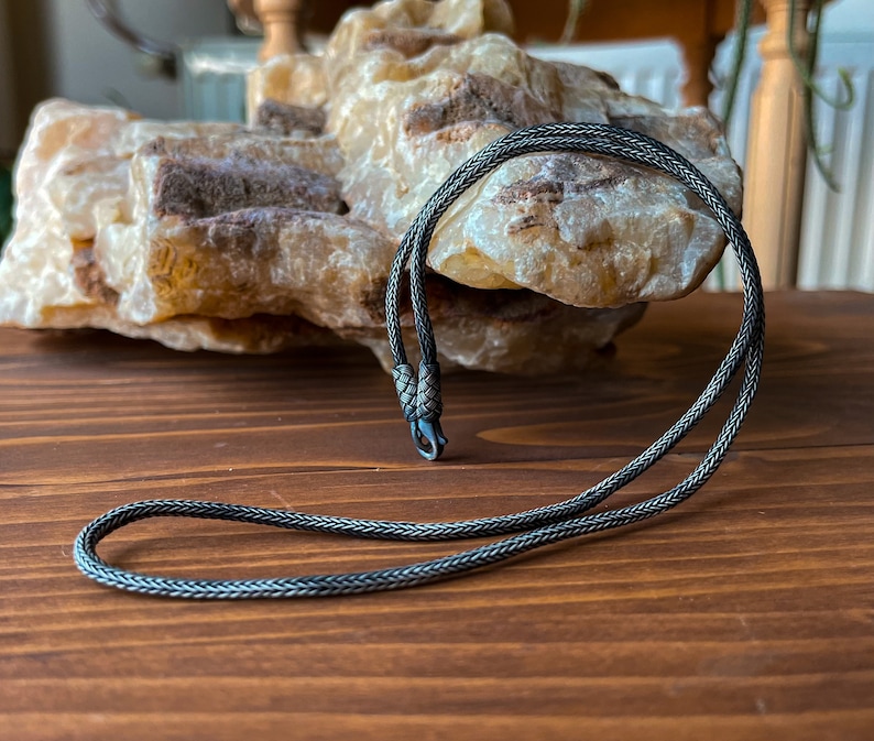Pode incluir: Um colar de corrente de prata com um design tran&ccedil;ado e um fecho. O colar est&aacute; sobre uma superf&iacute;cie de madeira.