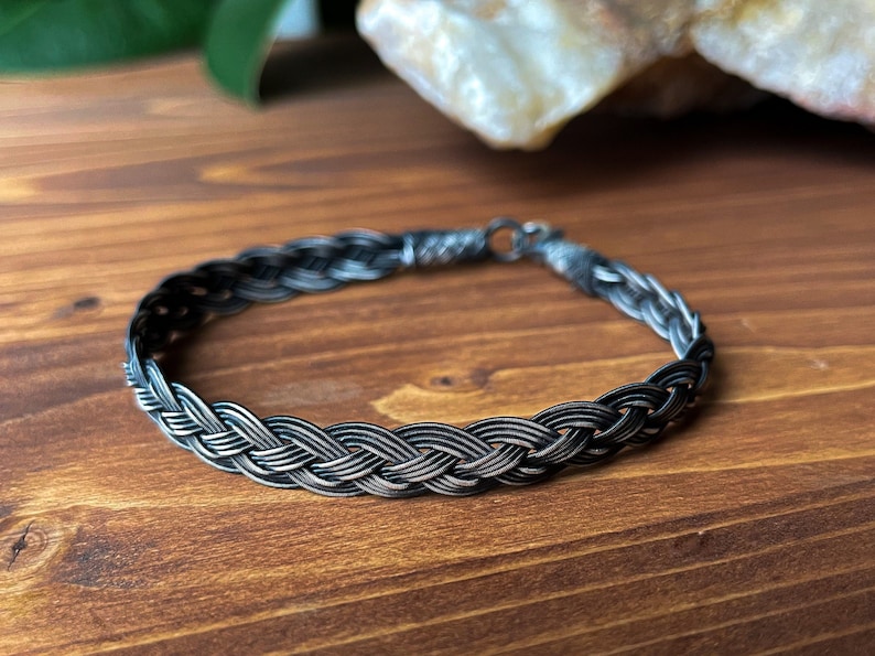 May include: A silver braided bracelet with a dark oxidised finish. The bracelet is made of multiple strands of silver wire that are woven together to create a braided design.