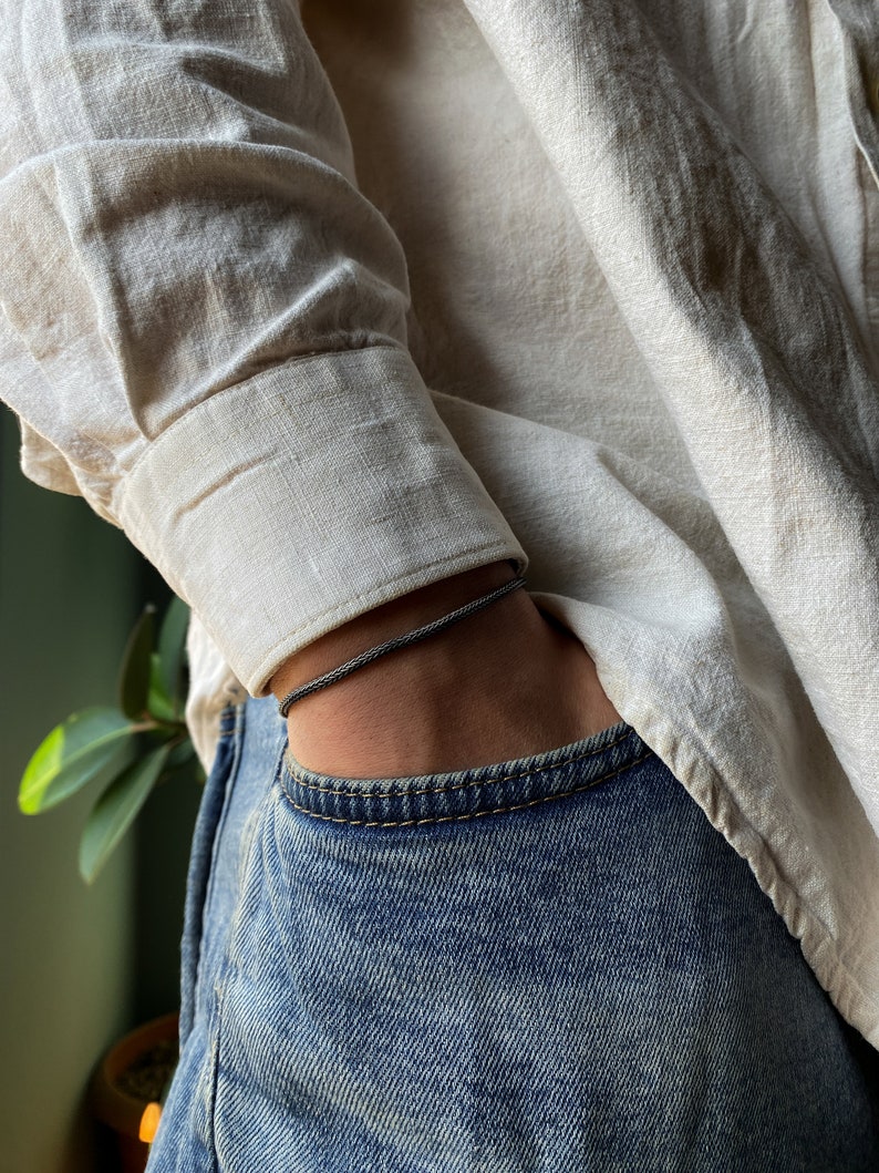 May include: A person wearing a light beige linen shirt with a dark gray bracelet on their wrist. The person is wearing blue jeans with a front pocket.