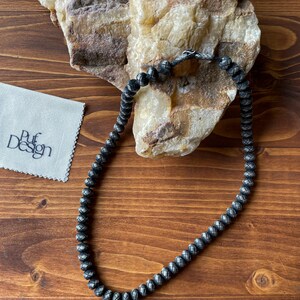 May include: A dark gray beaded necklace with a woven pattern, resting on a wooden surface. The necklace is next to a white cloth with the text "Puf Design". A large, light-colored rock is in the background.