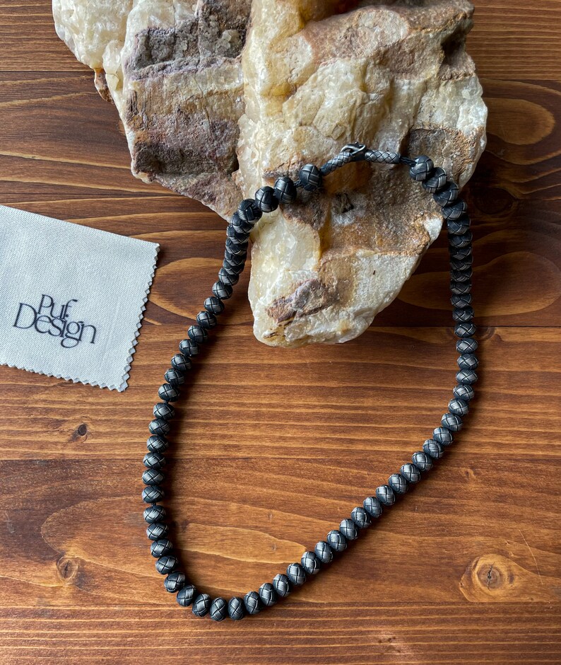 May include: A dark gray beaded necklace with a woven pattern, resting on a wooden surface. The necklace is next to a white cloth with the text "Puf Design". A large, light-colored rock is in the background.