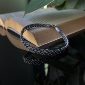 K&ouml;nnte beinhalten: Ein geflochtenes Silberarmband mit strukturiertem Design. Das Armband wird auf einer reflektierenden Oberfl&auml;che mit einem offenen Buch im Hintergrund pr&auml;sentiert. Das filigrane Geflecht des Armbands wird durch die warmen Farbt&ouml;ne der Buchseiten hervorgehoben.