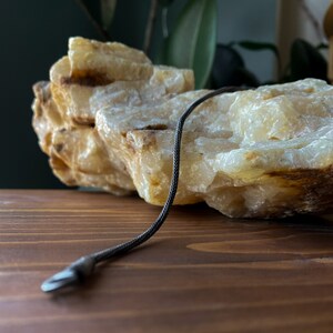 May include: A black braided cord with a silver metal clasp. The cord is lying on a light brown wooden surface next to a large, white, and yellow rock.