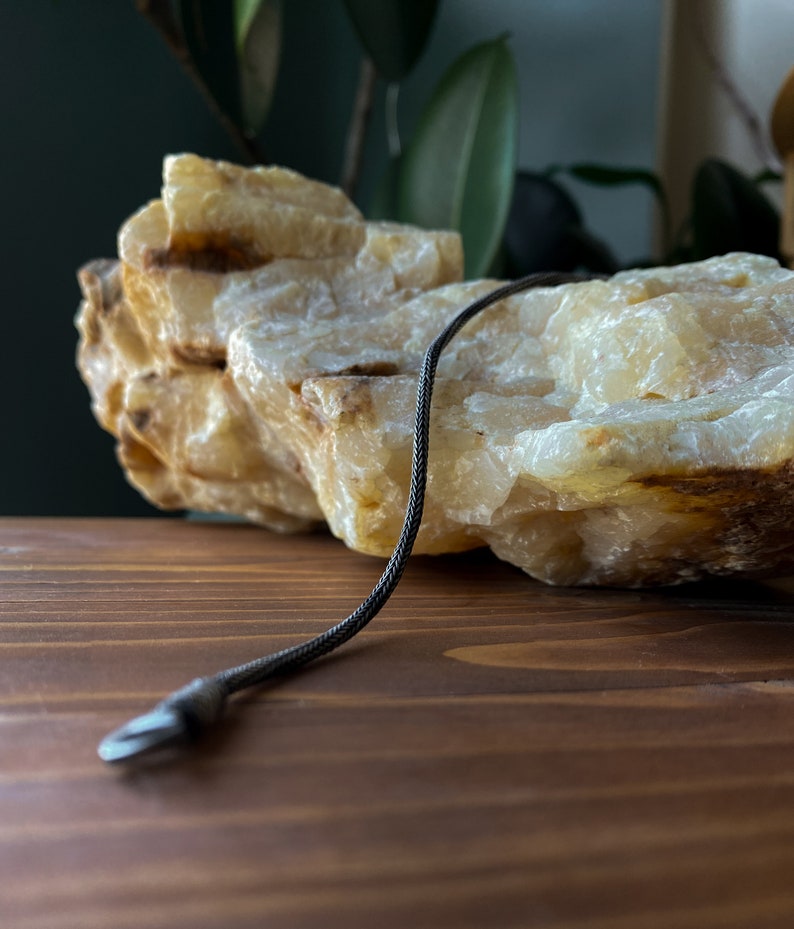 May include: A black braided cord with a silver metal clasp. The cord is lying on a light brown wooden surface next to a large, white, and yellow rock.