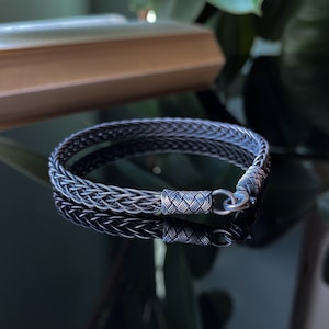 K&ouml;nnte beinhalten: Ein silbernes geflochtenes Armband mit einem gewebten Design. Das Armband hat einen Verschluss und wird auf einer reflektierenden Oberfl&auml;che gezeigt. Das komplizierte Muster und der metallische Glanz des Armbands werden hervorgehoben.