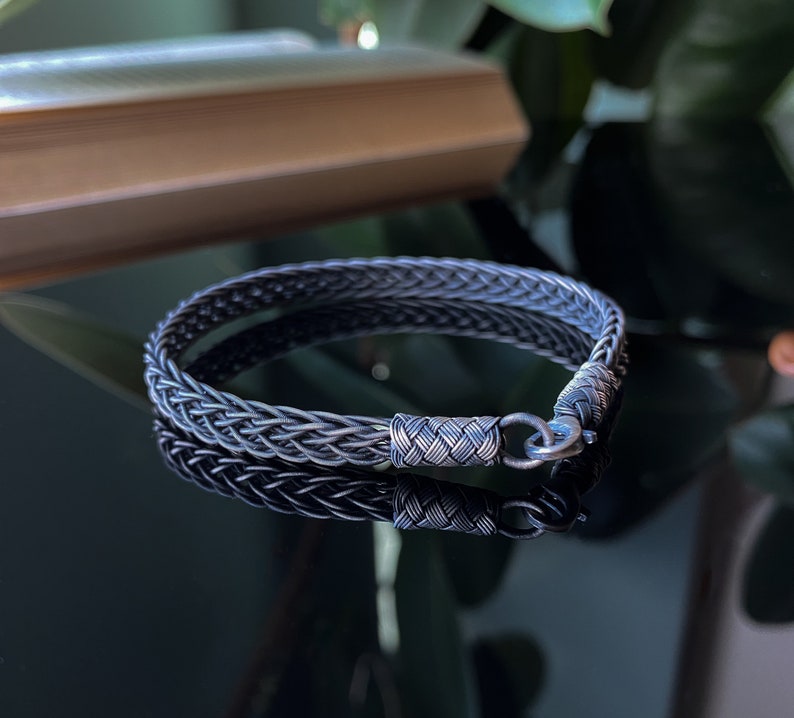 K&ouml;nnte beinhalten: Ein silbernes geflochtenes Armband mit einem gewebten Design. Das Armband hat einen Verschluss und wird auf einer reflektierenden Oberfl&auml;che gezeigt. Das komplizierte Muster und der metallische Glanz des Armbands werden hervorgehoben.