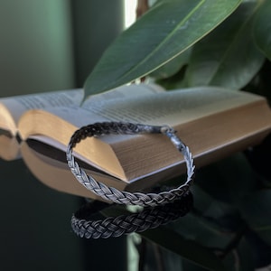 May include: A silver braided bracelet with a woven design. The bracelet is lying on a book with a green plant in the background.