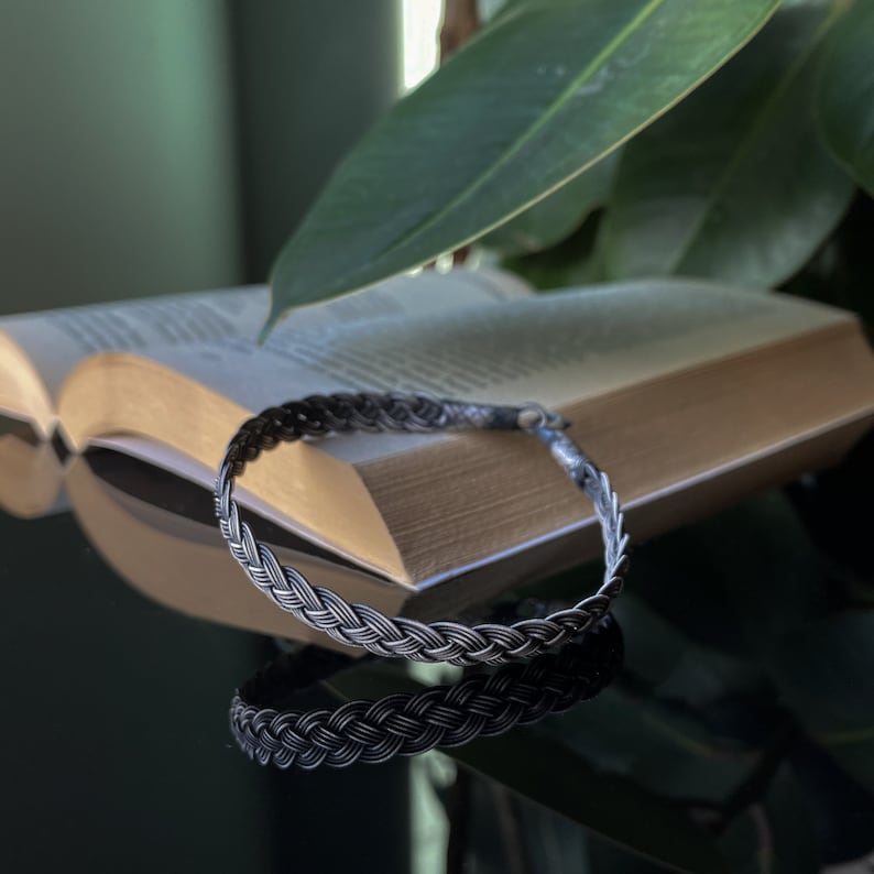 May include: A silver braided bracelet with a woven design. The bracelet is lying on a book with a green plant in the background.