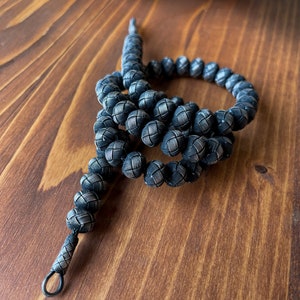 May include: A handcrafted, dark silver-toned beaded necklace. The beads are intricately woven, creating a textured, spherical design. The necklace is displayed on a wooden surface, showcasing the detailed craftsmanship of the jewelry.