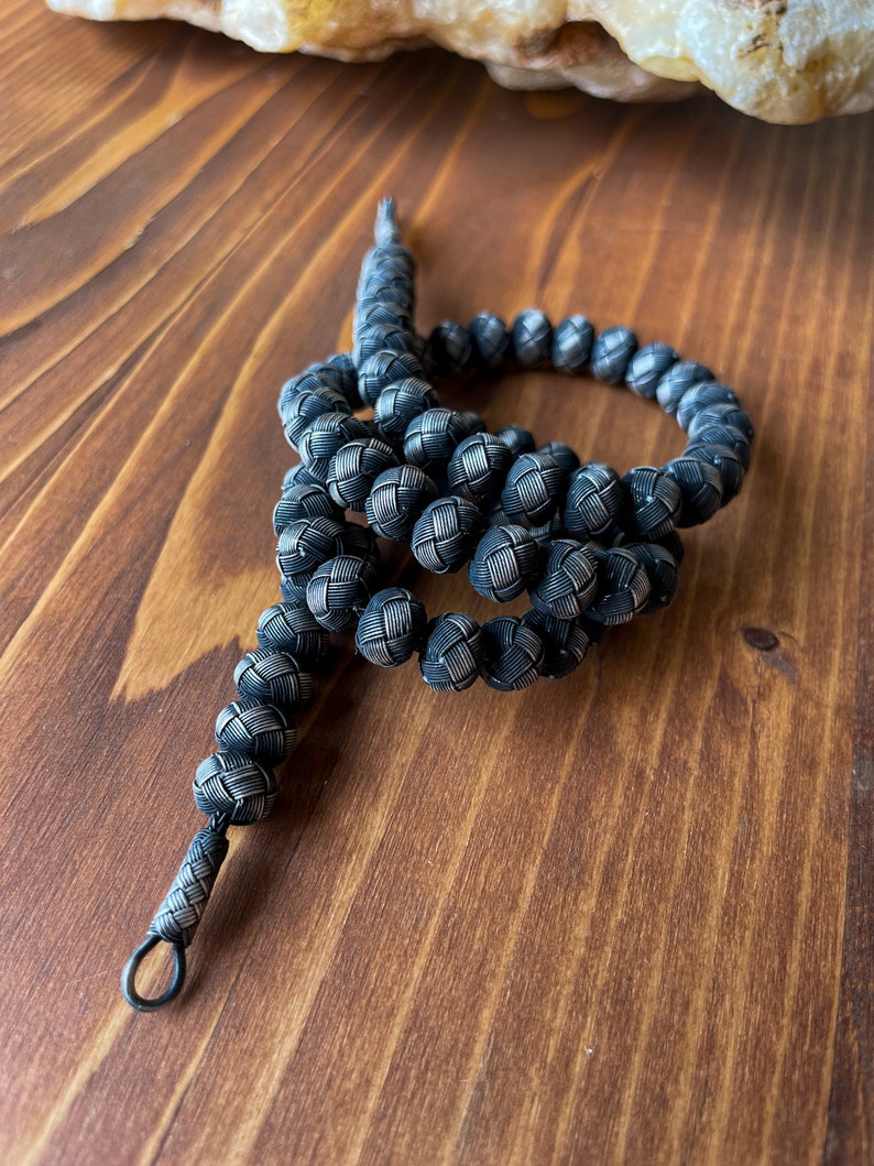May include: A handcrafted, dark silver-toned beaded necklace. The beads are intricately woven, creating a textured, spherical design. The necklace is displayed on a wooden surface, showcasing the detailed craftsmanship of the jewelry.