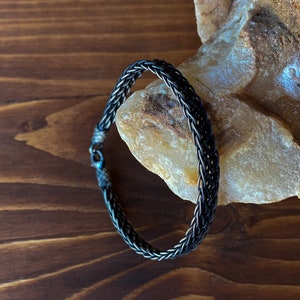 K&ouml;nnte beinhalten: Ein geflochtenes Silberarmband mit dunklem Finish. Das Armband liegt auf einem hellfarbigen Stein vor einer dunkelbraunen Holzoberfl&auml;che. Das Armband hat einen Verschluss.
