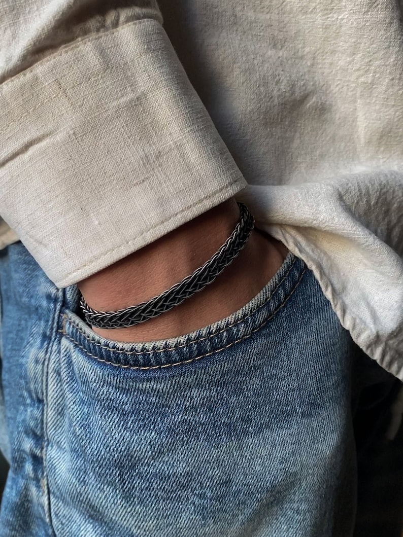 K&ouml;nnte beinhalten: Ein geflochtenes Silberarmband wird am Handgelenk getragen. Das Armband besteht aus miteinander verflochtenen Metallstr&auml;ngen, wodurch ein strukturiertes Aussehen entsteht. Die Person tr&auml;gt ein helles Hemd und eine blaue Jeans.