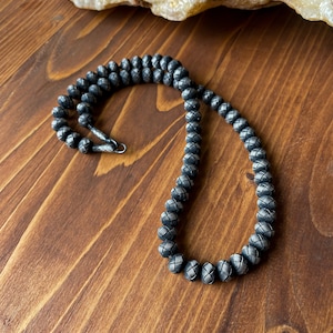 May include: A dark gray beaded necklace with a woven pattern. The necklace is made of small, round beads and has a silver clasp. The necklace is displayed on a wooden surface.