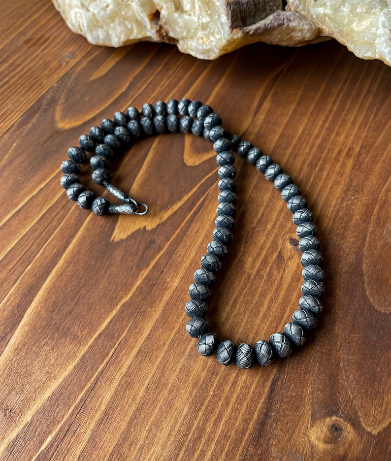 May include: A dark gray beaded necklace with a woven pattern. The necklace is made of small, round beads and has a silver clasp. The necklace is displayed on a wooden surface.