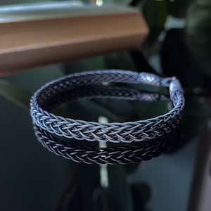 K&ouml;nnte beinhalten: Ein geflochtenes Silberarmband mit einem strukturierten, gewebten Design. Das Armband ist kreisf&ouml;rmig und spiegelt sich auf einer dunklen, gl&auml;nzenden Oberfl&auml;che. Der Verschluss ist am Ende des Armbands sichtbar. Es ist ein Schmuckst&uuml;ck.