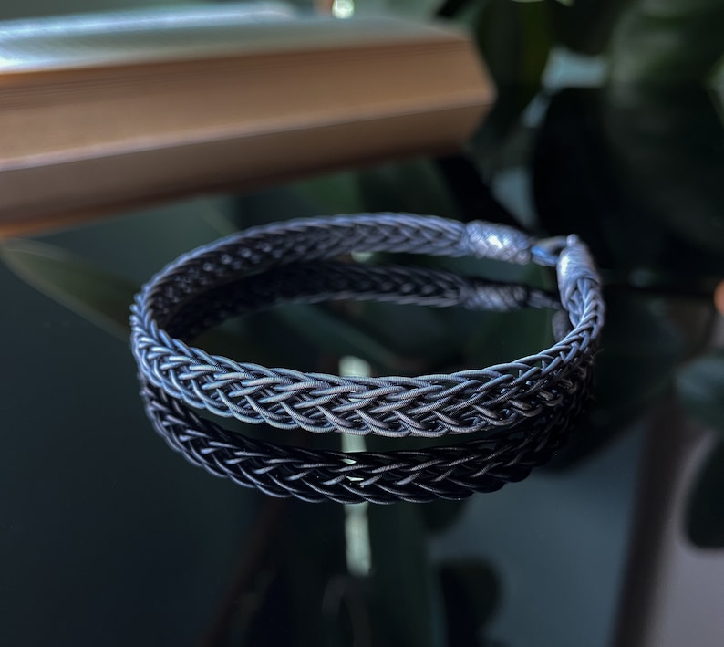 K&ouml;nnte beinhalten: Ein geflochtenes Silberarmband mit einem strukturierten, gewebten Design. Das Armband ist kreisf&ouml;rmig und spiegelt sich auf einer dunklen, gl&auml;nzenden Oberfl&auml;che. Der Verschluss ist am Ende des Armbands sichtbar. Es ist ein Schmuckst&uuml;ck.