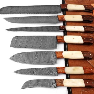 May include: A set of seven kitchen knives with Damascus steel blades featuring a wavy pattern, wooden handles with white bone inlays, and brass pins. The knives are arranged horizontally next to a brown leather roll with metal snap fasteners. F-15.