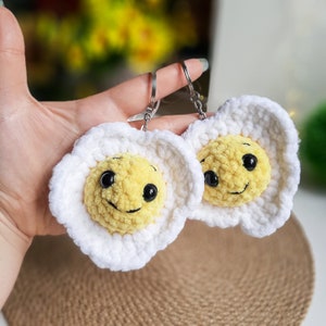 Puede incluir: Dos llaveros de ganchillo en forma de huevo con caras sonrientes. Los llaveros son blancos con hilo amarillo para la yema del huevo e hilo negro para los ojos y la boca.