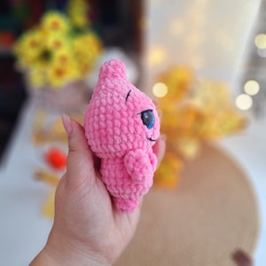 May include: A pink crocheted plush toy with blue eyes. The toy is shaped like a teardrop and has a small smile.