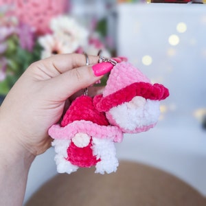 May include: Two pink and white yarn gnome keychains with a silver key ring. The gnomes have pink hats and white beards.