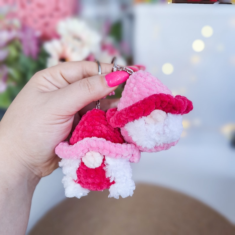 May include: Two pink and white yarn gnome keyrings with a silver key ring. The gnomes have pink hats and white beards.