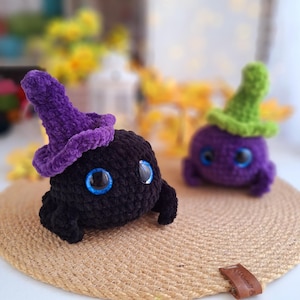 May include: Two crocheted plush toys shaped like round creatures with large eyes. One is black with a purple hat and the other is purple with a green hat.