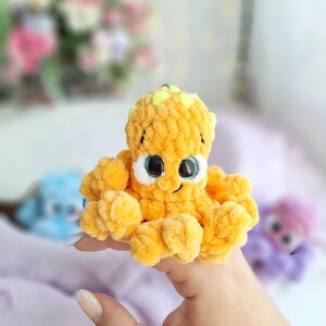 May include: A handmade, plush orange octopus toy with large eyes and a smiling mouth. The octopus has a textured, fuzzy appearance. Other similar plush toys are visible in the background, suggesting a collection or set of toys.