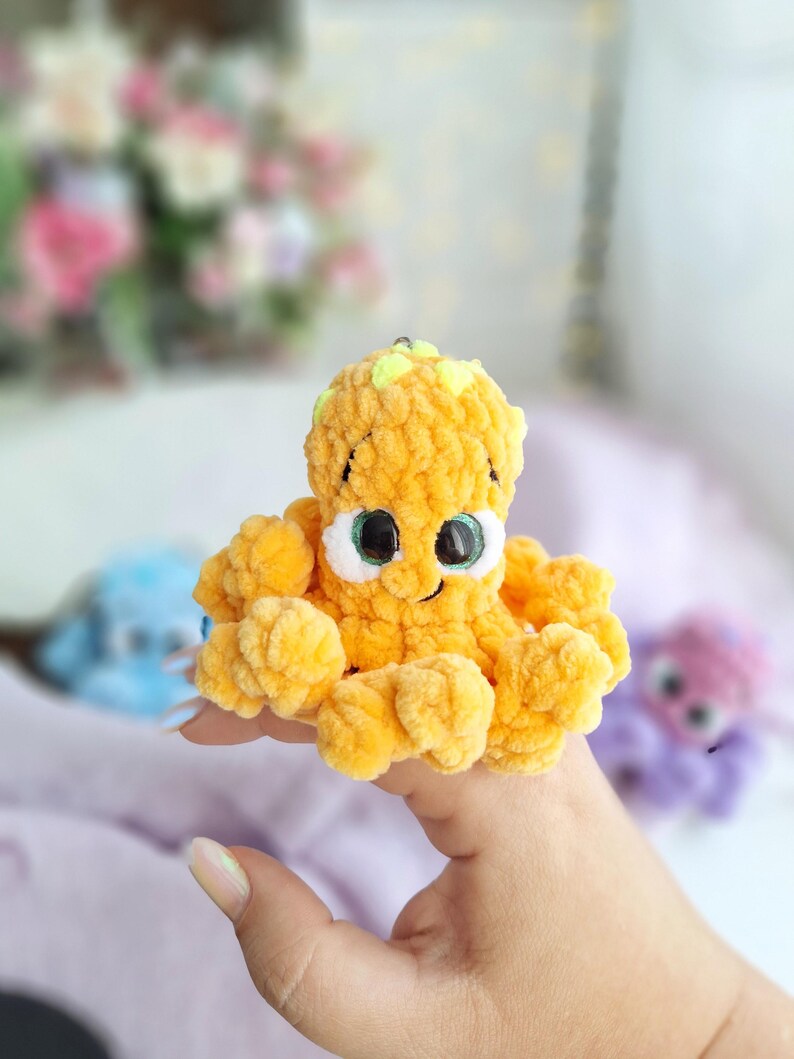 May include: A handmade, plush orange octopus toy with large eyes and a smiling mouth. The octopus has a textured, fuzzy appearance. Other similar plush toys are visible in the background, suggesting a collection or set of toys.
