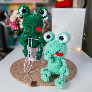 May include: Two plush frog toys in the image. One is green and seated in a white wire basket, the other is light green. Both frogs have large, round eyes with white and black details, and red tongues sticking out.