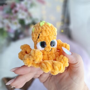 May include: A small, handmade orange crochet octopus toy with a smiling face and large eyes. The octopus has eight curly tentacles and a small green accent on its head. The plush toy is soft and cuddly, ideal for a child's toy collection.