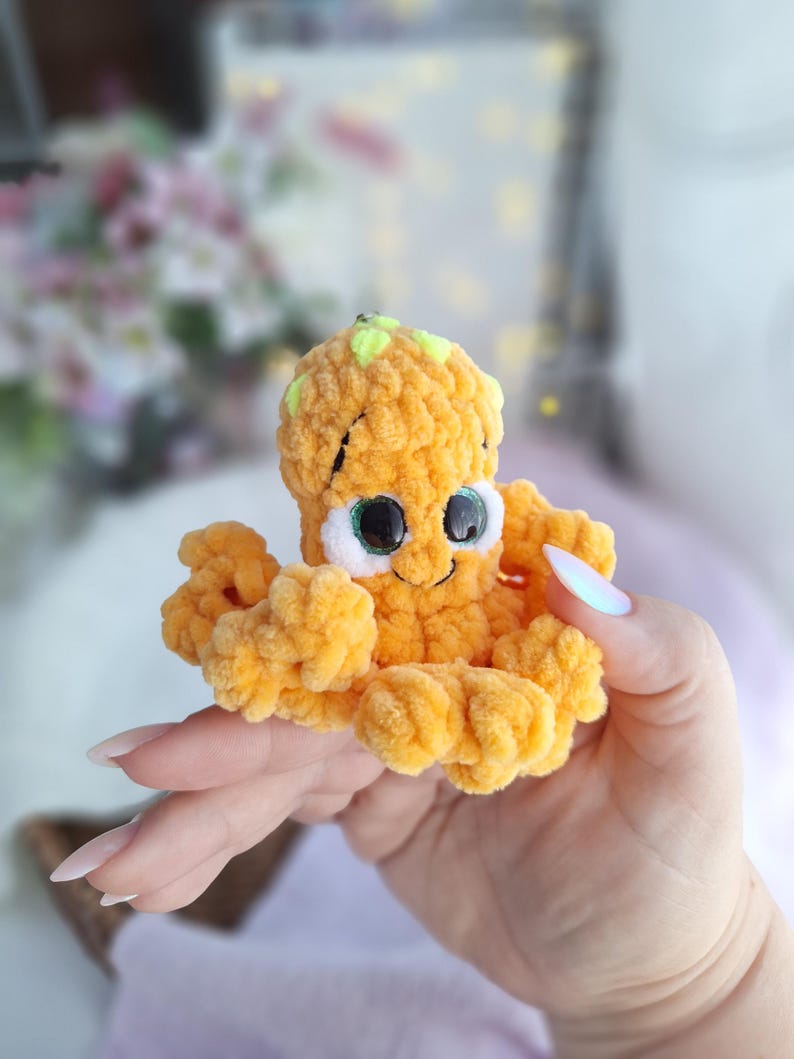 May include: A small, handmade orange crochet octopus toy with a smiling face and large eyes. The octopus has eight curly tentacles and a small green accent on its head. The plush toy is soft and cuddly, ideal for a child's toy collection.