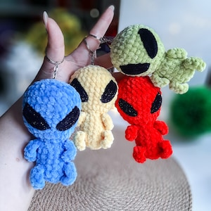 Może przedstawiać: Cztery breloczki z postaciami kosmitów wykonane na szydełku w kolorze niebieskim, żółtym, czerwonym i zielonym. Każdy breloczek ma czarne oko i uśmiechnięte usta.