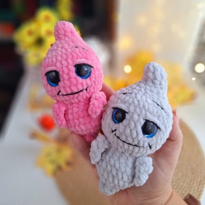 May include: Two crocheted plush ghost toys, one pink and one gray, with blue eyes and a smiling mouth. The toys are made of soft yarn and have a whimsical, friendly appearance.
