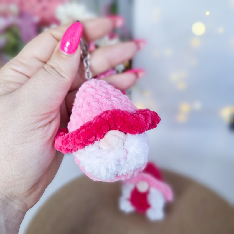 May include: A pink and white crocheted gnome keychain with a pink hat and white beard. The gnome is holding a small pink heart.