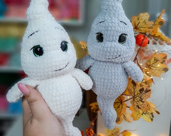 May include: Two crocheted ghost plushies, one white and one gray, with green eyes and a smiling expression. The plushies are sitting on a bed of fall leaves.