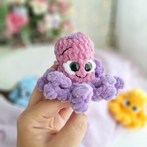 May include: A crocheted octopus toy in pink and purple. The toy has large, googly eyes and a smiling face. The tentacles are a soft purple. The toy is held in a hand, with other similar toys visible in the background.