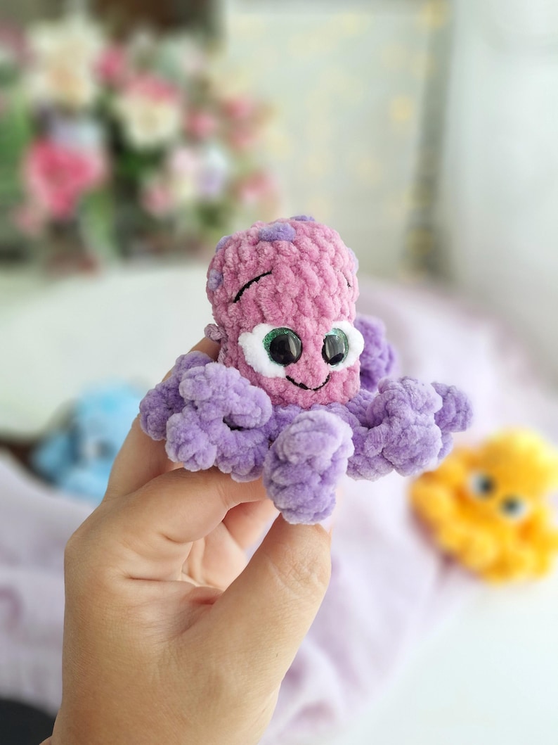 May include: A crocheted octopus toy in pink and purple. The toy has large, googly eyes and a smiling face. The tentacles are a soft purple. The toy is held in a hand, with other similar toys visible in the background.
