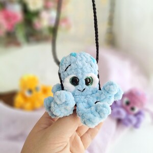 May include: A light blue crocheted octopus pendant on a black cord. The octopus features large eyes and small blue spots. Other colourful crocheted octopuses are visible in the background.