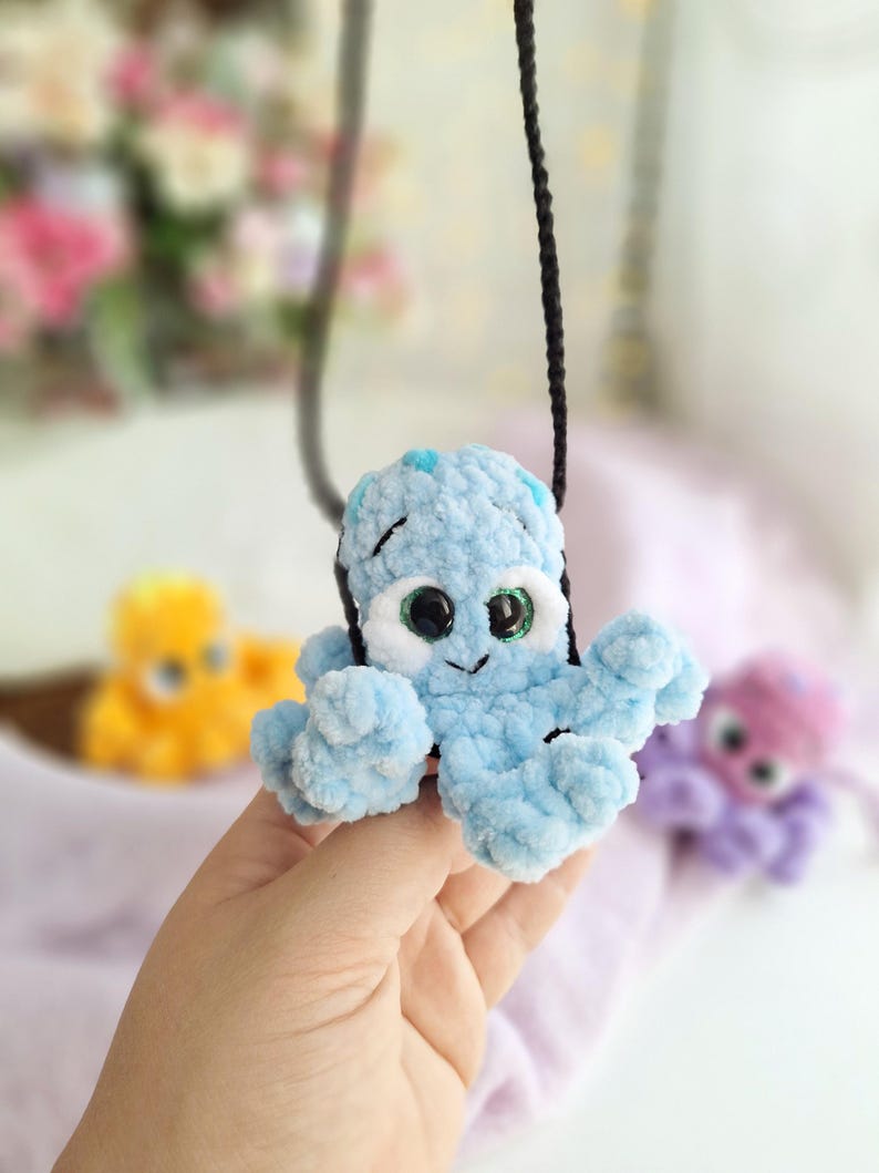 May include: A light blue crocheted octopus pendant on a black cord. The octopus features large eyes and small blue spots. Other colourful crocheted octopuses are visible in the background.