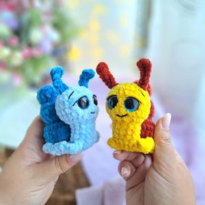 May include: Two handmade crochet snail toys. One is blue with darker blue accents, the other is yellow with red accents. Both have large, expressive eyes and smiling faces. The toys are held in hands, suggesting a small size, perfect for children.