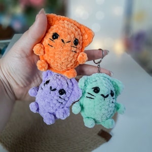 May include: Three crocheted cat keychains in orange, purple, and green. The keychains are attached to a silver metal ring.