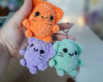 Peut inclure: Trois porte-clés en forme de chat en crochet de couleur orange, violet et vert. Les porte-clés sont attachés à un anneau en métal argenté.